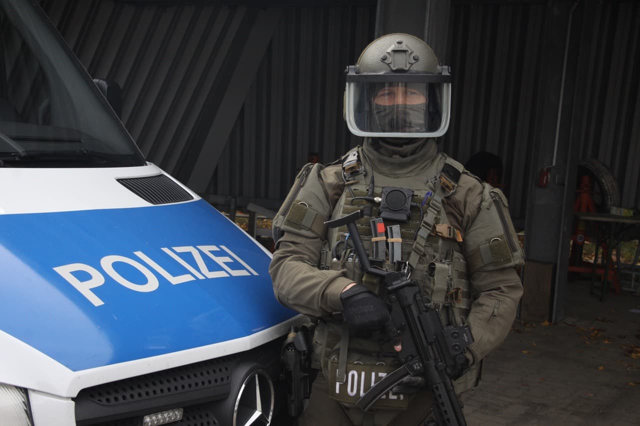 SEK-Operator in Einsatzmontur mit Helm, Plattenträger und Maschinenpistole vor Polizeifahrzeug